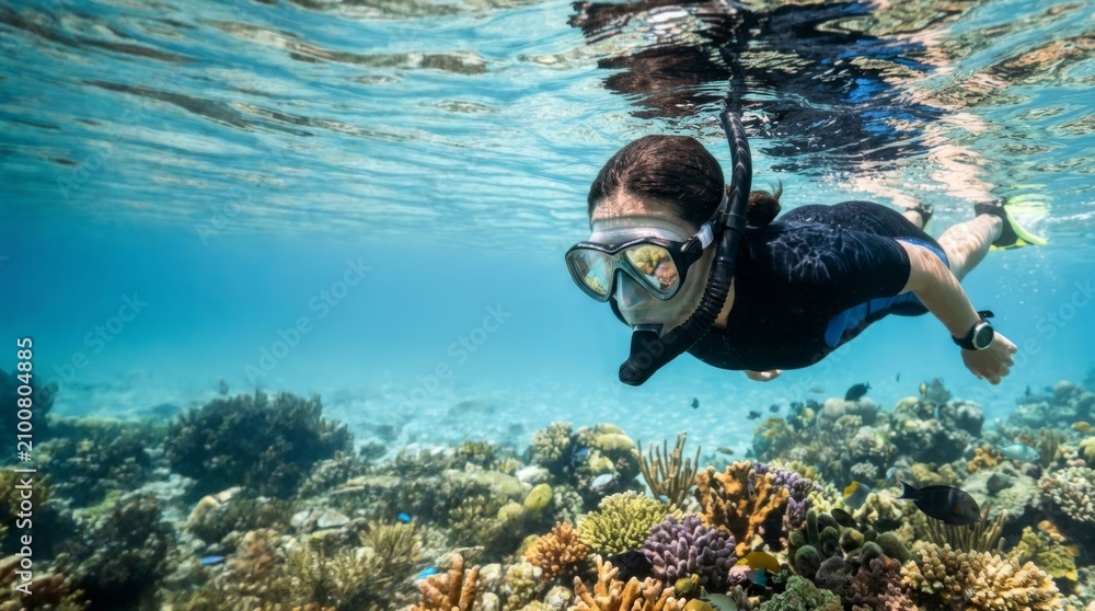 Obraz premium Woman snorkeling over vibrant coral reef and tropical fish, clear turquoise water with sailboat silhouette, active vacation lifestyle