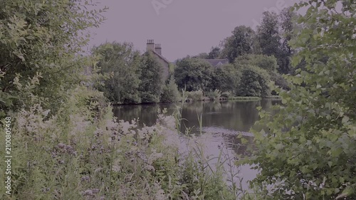 Picturesque tranquil scene of part of a village pond viewed between trees with a house with tall chimneys in the background. No location markers or people.

