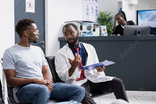 Tableau sur toile Professional doctor with clipboard talks with male patient seated on clinical lobby bench while general practitioner offers supportive medical advice