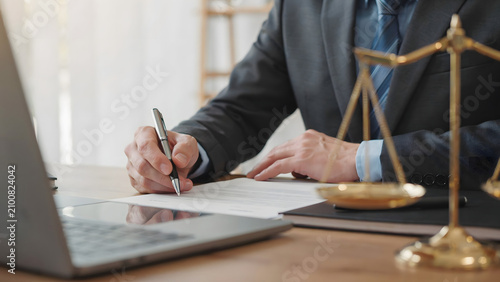 A lawyer in a dark suit sitting at a wooden desk, writing on a legal document with a metal pen, with a golden scale of justice nearby
