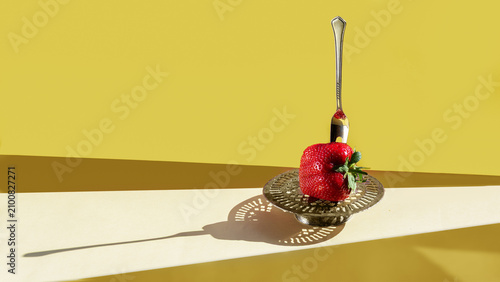 Conceptual food art composition featuring a single fresh strawberry pierced by a vintage silver dessert fork on a yellow background.