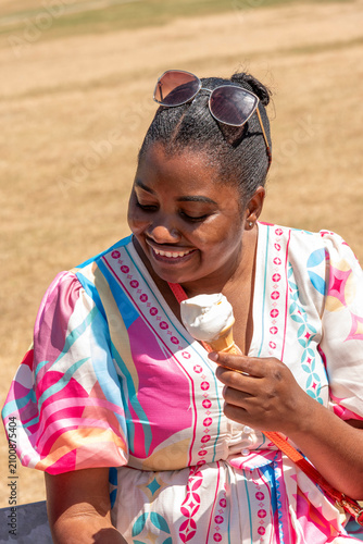 Nottingham England UK. 11.07.2026. A smiling woman enjoys a cold treat on a warm day, wearing a colorful patterned dress and sunglasses perched on her head