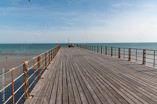 Bognor Regis  West Sussex England UK. 14.07.2026. A weathered wooden pier stretches towards the calm sea under a clear blue sky, with a sandy beach and holidaymakers visible in the distance