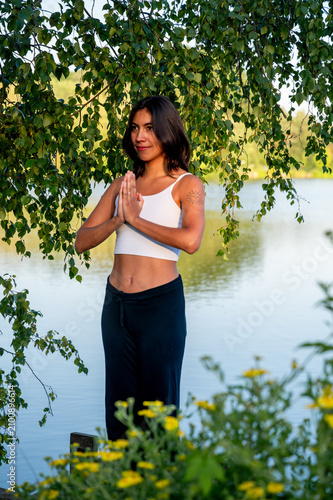 A young Hispanic woman practises the chair pose with prayer hands in a green park beside a calm lake at sunset, building strength, flexibility and focus in the fresh air.