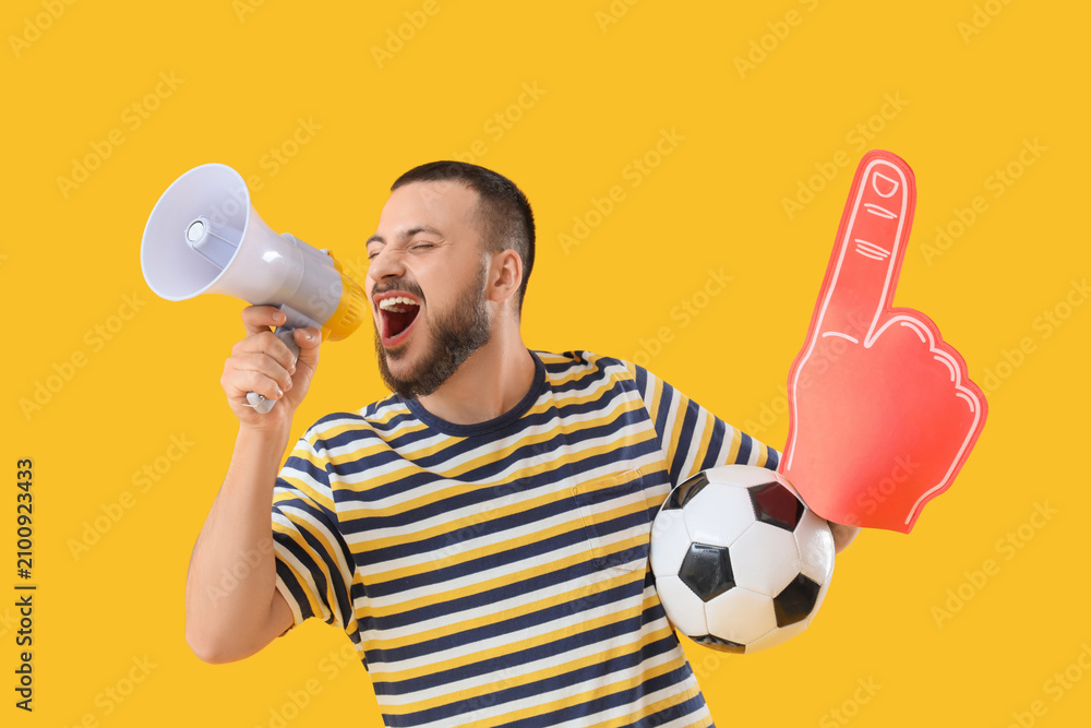 Obraz premium Male sports fan with soccer ball and foam hand shouting into megaphone on yellow background