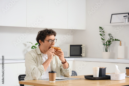 Young man eating tasty hot dog at table in kitchen