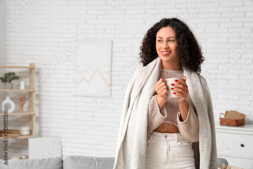 Beautiful young African-American woman with cozy blanket and cup of tea at home