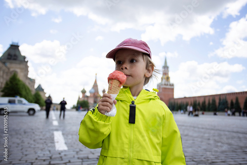 A happy four-year-old child eating strawberry ice cream in a waffle cone on Red Square in Moscow, Russia. The child is wearing a bright neon green jacket. A colorful travel and lifestyle 