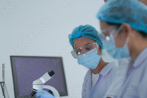 Fotografie Two lab colleagues inspects a laboratory sample with focus before monitors showing genetic data while wearing white lab coats and blue protective gloves and modern scientific innovation