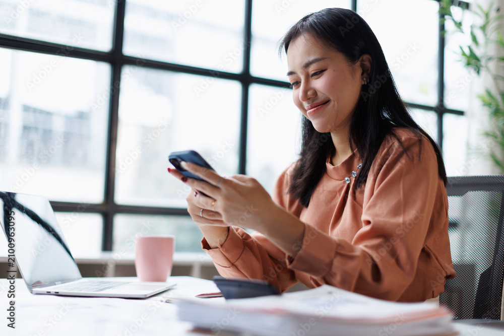 Obraz premium Young asian businesswoman sitting at office desk, messaging on smartphone while working, managing tasks, using technology for communication and remote work