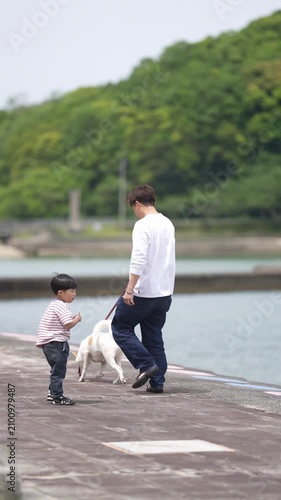 Man walks child and dog along waterfront path. Child walks beside him, holding leash. Dog trots happily beside them both. Trees line the water's edge behind. Perfect for family or pet outdoor moments