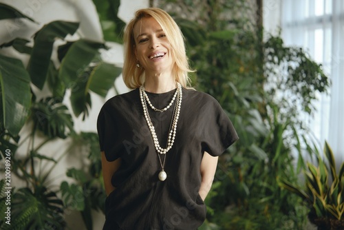 A cheerful woman with blonde hair poses confidently amidst a backdrop of vibrant plants in a bright indoor area. She wears a stylish outfit with layered necklaces, exuding a sense of warmth and joy