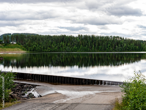 Obraz na płótnie Calm reservoir and overflow dam at Spegeldammen in Vuollerim, northern Sweden, reflecting a dense boreal forest beneath a dramatic cloudy sky