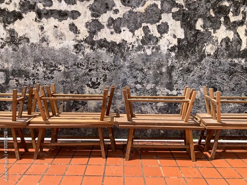 Rows of long wooden benches neatly stored on a brown tiled floor beside a weathered school wall under bright natural sunlight