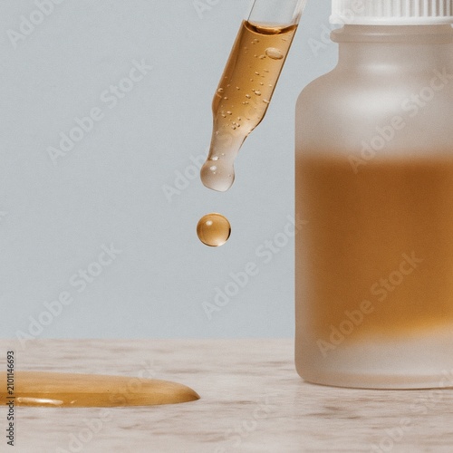 A golden serum drop falling from a glass pipette onto a marble surface, showing a viscous honey texture for clean beauty and skincare advertising