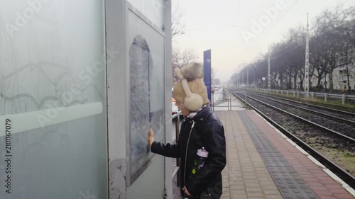 Outdoor medium shot captures a young girl in winter clothes making a funny face while wiping condensation off a foggy city map pane at an urban tram stop alongside empty railway tracks in winter.