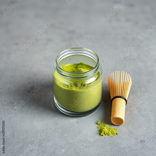 Glass Jar of Ceremonial Green Matcha Powder with Traditional Bamboo Whisk on Gray Stone Surface with Copy Space