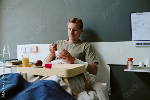 Obraz na plátně Young adult Caucasian man recovering in military hospital bed, eating meal during treatment and convalescence