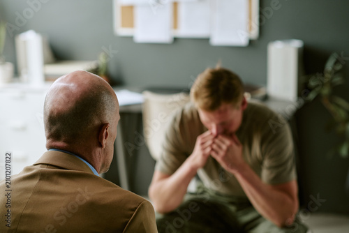 Young adult Caucasian man sitting in military hospital, covering face and receiving counseling support from clinician, conveying trauma recovery. Ideal for veteran mental health campaigns