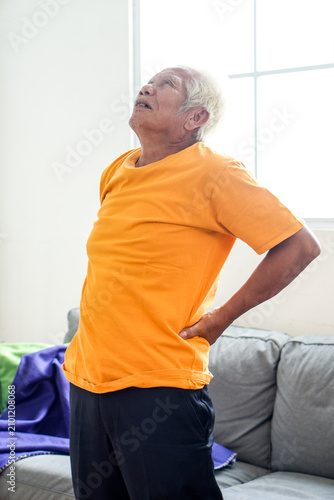 Senior Asian Man Holding His Lower Back in Pain While Standing by Sofa at Home