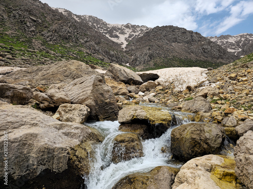 Obraz na plátně A rocky mountain stream cascades over boulders and mossy rocks near a snow cave opening, with patches of avalanche snow and rugged snow-capped peaks rising behind green hillsides