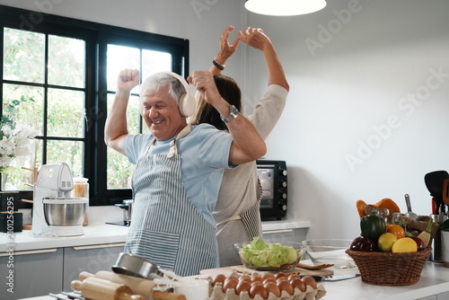 An elderly couple, a grandfather wearing headphones and a grandmother in an apron, bake bread and dance together at the kitchen counter in their home