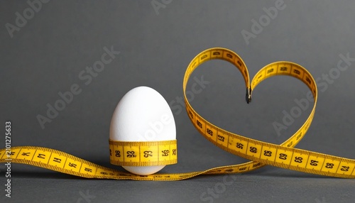 White egg wrapped in yellow measuring tape formed into heart shape on gray background with measuring nutrition calories