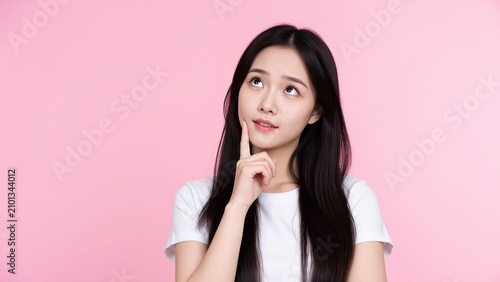 Pensive Young Woman: A young woman with long dark hair, wearing a white t-shirt, rests her chin on her hand, gazing upwards with a thoughtful expression against a soft pink background.