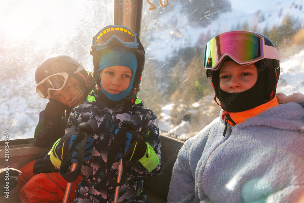 Obraz premium Children of different ages ride ski gondola in winter gear, with bored teenager leaning by window while younger skiers look cheerful. Warm sunlight, helmets, goggles and poles frame family ski trip