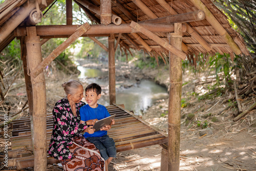 Elderly woman and young boy sit together on bamboo platform under rustic shelter beside quiet stream, smiling while sharing tablet in sunlit rural setting with natural textures, gentle greenery, and