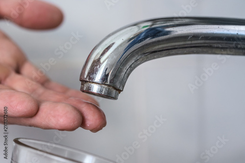 Adult hand held under chrome faucet spout with corroded aerator and mineral deposits, dry plumbing fixture showing no water flow, soft neutral background, natural skin tones and detailed metal surface