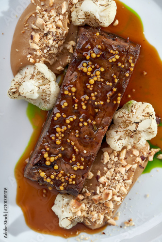 Macro shot of roasted meat glazed with mustard seeds, served with cauliflower and gravy on a white plate. Elegant fine dining close-up.