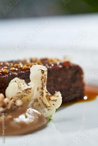 Macro shot of roasted meat glazed with mustard seeds, served with cauliflower and gravy on a white plate. Elegant fine dining close-up.