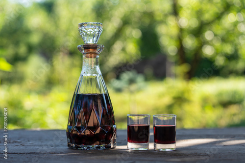 Homemade cherry liqueur in decanter and two shot glasses. Crystal carafe filled with dark red cherry liqueur next two filled shot glasses on rustic wooden table on green nature background