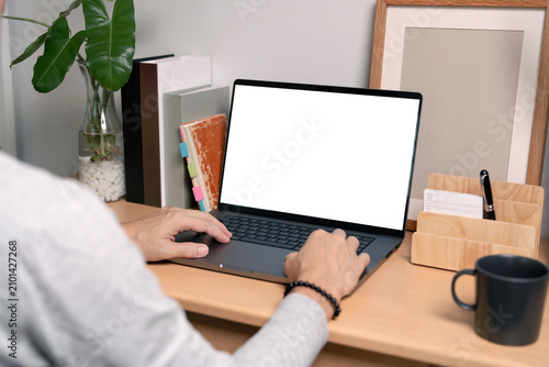 Man working on laptop with blank mockup screen on wooden desk, over the shoulder view.