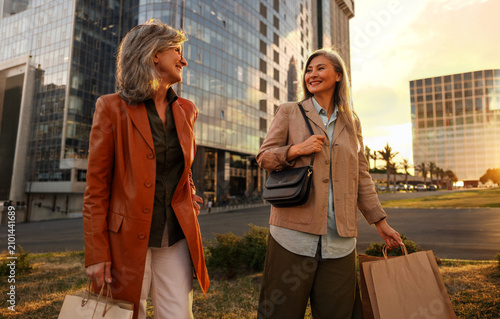 Cuadro en lienzo Two mature women, one in a rust orange blazer and one in a tan jacket carrying p