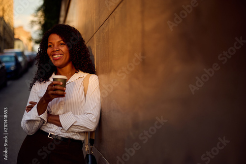Smiling African American Woman With Coffee Leaning On City Wall for Casual Business and Urban Lifestyle