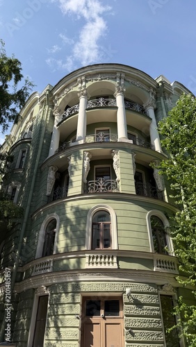 Historic green building facade with white columns, elegant caryatid statues supporting curved wrought-iron balconies, and arched windows framed by green trees under a blue summer sky.