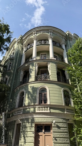 Bild auf Leinwand Elegant rounded corner of a historic green building featuring white columns, caryatid sculptures, intricate balconies, and arched windows surrounded by lush green foliage on a sunny day