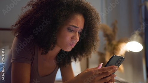 Frustrated African American woman using mobile phone losing bet online failure in bed at night disappointed ethnic girl female scrolling smartphone lose internet betting user upset lying in bedroom