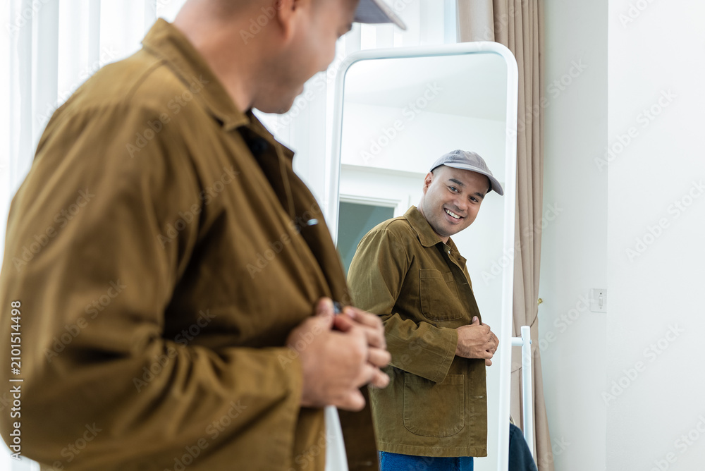 Fototapeta premium Smiling Asian man buttoning his casual brown jacket in front of a mirror. Concept of morning routine preparation, trendy street fashion and everyday confidence.