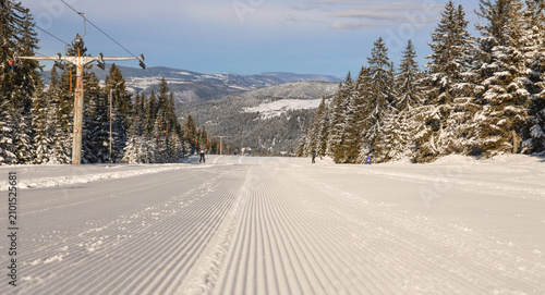 Canvastavla Freshly groomed ski slope with corduroy snow, distant skiers, chairlift infrastructure and snowy mountain scenery on a sunny winter day