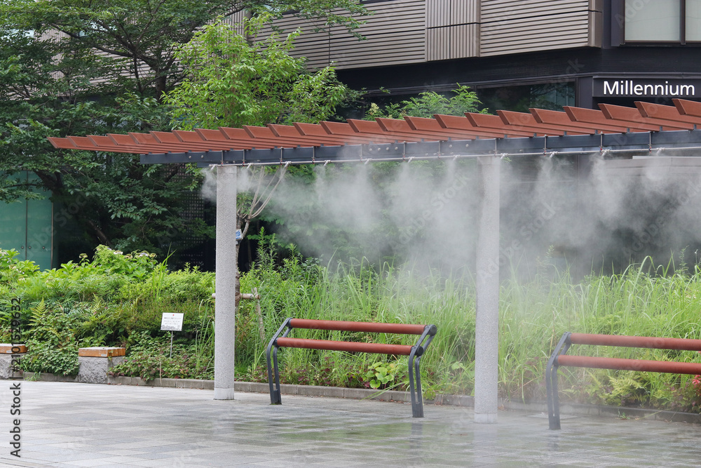 Obraz premium TOKYO, JAPAN - July 16, 2026: Cooling mist emitted from a device over benches in Ginza on a hot summer's day. The Millennium Mitsui Garden Hotel behind it.