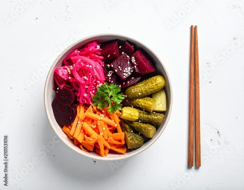 Top view of a ceramic bowl filled with colorful pickled vegetables and chopsticks on a white surface
