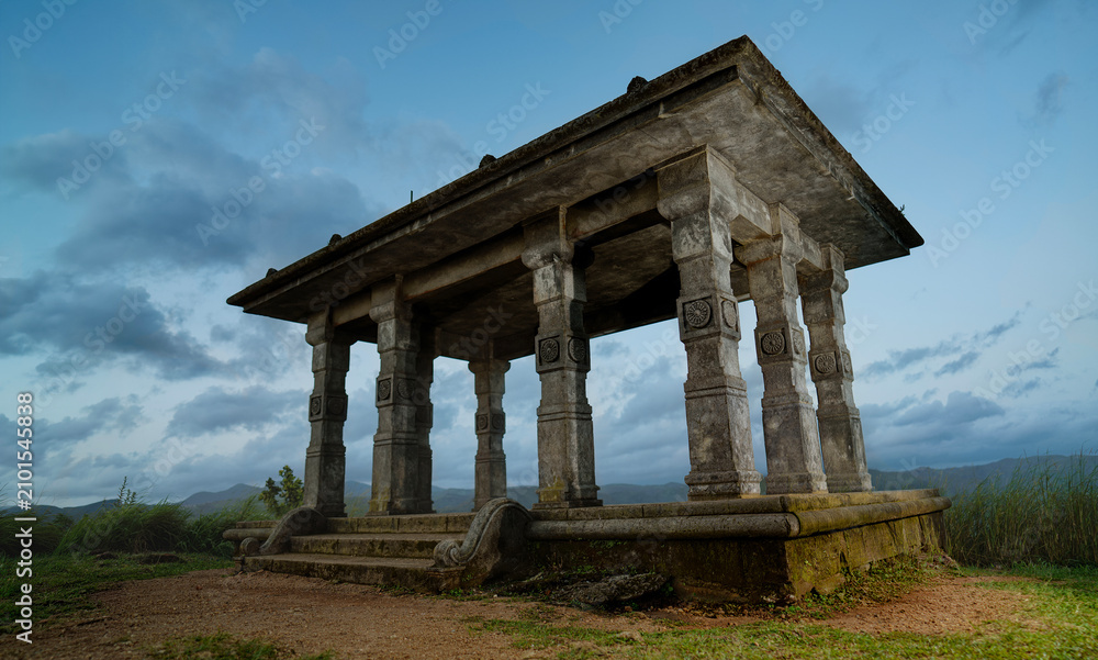 Obraz premium An ancient stone pavilion with carved columns stands on a grassy hilltop, framed by dramatic clouds and distant mountains at dusk, conveying atmosphere, solitude, heritage and weathered craftsmanship.