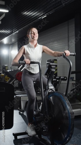Vertical video of young Caucasian woman exercising on air bike in gym. Footage for fitness brand commercials, gym promotional videos, and wellness campaigns