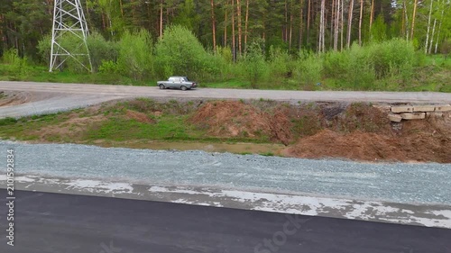 A parallel flight of stairs behind a car driving along a forest road alongside an asphalt road under construction in a pine forest on a sunny summer evening. Eastern Bypass, Novosibirsk, Russia.