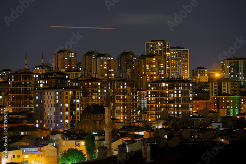 View of typical turkish multistory residential buildings with illuminated windows, warm street lighting and airplane light trail in Istanbul, Turkey at night. Cityscape and urban concept