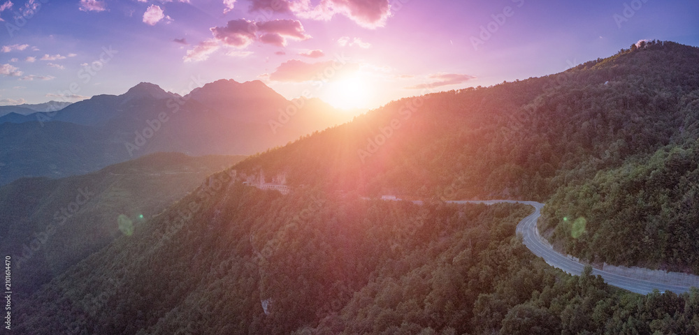 Obraz premium Winding mountain road traversing a lush green forest with dramatic sun flare and mountain peaks during a vibrant sunset