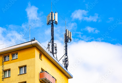 Mobile cellular network antennas and telecommunication transmitters installed on the roof of a yellow residential apartment building against blue sky